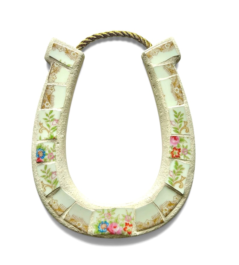 May include: A decorative horseshoe made of mosaic tiles with floral patterns and gold accents. The horseshoe is cream-colored with a gold rope for hanging. The mosaic tiles feature pink, blue, and red flowers.