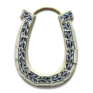 May include: A horseshoe-shaped wall hanging. The horseshoe is made of white tiles with a blue leaf design. A gold-colored rope is attached at the top for hanging. The horseshoe is a symbol of good luck.