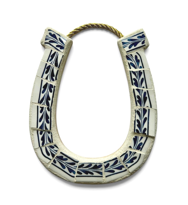 May include: A horseshoe-shaped wall hanging. The horseshoe is made of white tiles with a blue leaf design. A gold-colored rope is attached at the top for hanging. The horseshoe is a symbol of good luck.