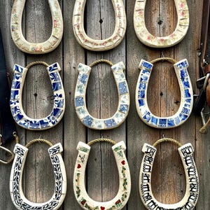 May include: Nine decorative horseshoe-shaped wall hangings. Each horseshoe is adorned with a unique mosaic design, featuring floral patterns, blue and white tiles, and other decorative elements. They hang against a weathered wooden background.