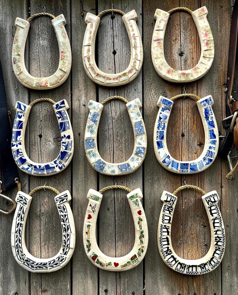 May include: Nine decorative horseshoe-shaped wall hangings. Each horseshoe is adorned with a unique mosaic design, featuring floral patterns, blue and white tiles, and other decorative elements. They hang against a weathered wooden background.