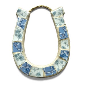 May include: A decorative horseshoe made of mosaic tiles. The tiles are a mix of blue and white with floral and ribbon patterns. A gold-colored rope is attached at the top for hanging. The horseshoe is a symbol of good luck.