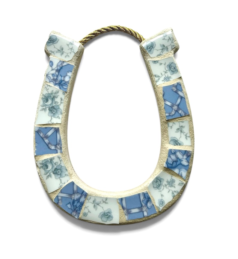 May include: A decorative horseshoe made of mosaic tiles. The tiles are a mix of blue and white with floral and ribbon patterns. A gold-colored rope is attached at the top for hanging. The horseshoe is a symbol of good luck.