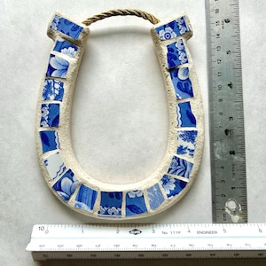 May include: A ceramic horseshoe with a mosaic design. The horseshoe is cream-colored with blue and white floral patterned tiles. A twisted rope is attached at the top for hanging. The horseshoe measures approximately 6 inches in height.