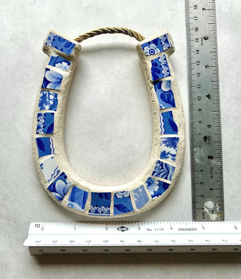 May include: A ceramic horseshoe with a mosaic design. The horseshoe is cream-colored with blue and white floral patterned tiles. A twisted rope is attached at the top for hanging. The horseshoe measures approximately 6 inches in height.