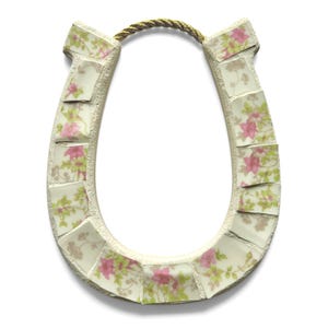 May include: A horseshoe-shaped decorative item with a mosaic design. The mosaic is made of small, irregular tiles with a floral pattern in pink and green on a cream background. A gold-colored rope is attached at the top.