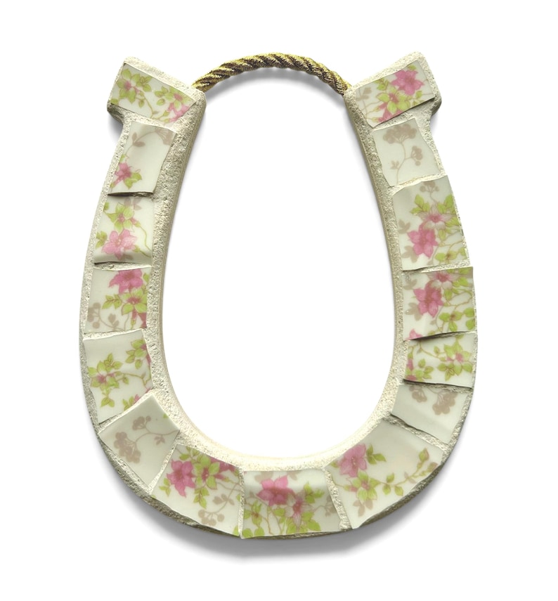 May include: A horseshoe-shaped decorative item with a mosaic design. The mosaic is made of small, irregular tiles with a floral pattern in pink and green on a cream background. A gold-colored rope is attached at the top.