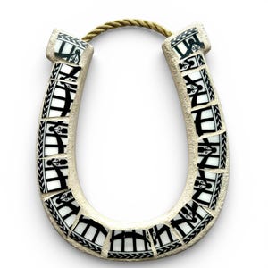 May include: A ceramic horseshoe with a mosaic design. The horseshoe is off-white with black and white mosaic tiles. A gold-colored rope is attached at the top. The horseshoe is a decorative item.