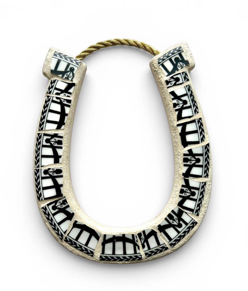 May include: A ceramic horseshoe with a mosaic design. The horseshoe is off-white with black and white mosaic tiles. A gold-colored rope is attached at the top. The horseshoe is a decorative item.
