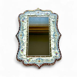 May include: A decorative rectangular mirror with a mosaic tile frame. The frame features a blue, white, and red floral design. The mirror's shape is ornate, with a scalloped top and sides. The mosaic tiles are a mix of broken pieces and whole tiles.