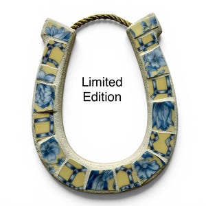 May include: A horseshoe-shaped wall hanging with a mosaic design. The mosaic features blue floral patterns on yellow tiles, set within a light-colored frame. The text "Limited Edition" is displayed on the front. A gold rope is attached at the top.
