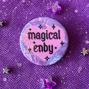 May include: A pink heart-shaped button with the text "magical enby" in black gothic font. The button has a holographic background with silver glitter stars.