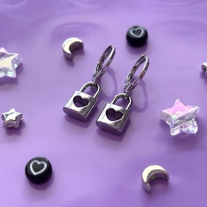 Peut inclure: Boucles d'oreilles cadenas argentées avec découpes en forme de cœur, accompagnées de breloques étoiles et lunes, et de perles noires avec des cœurs. L'ensemble est disposé sur une surface violette.
