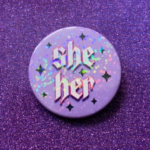 May include: A purple button with a glittery surface and black stars. The button says "she her" in white letters with a gothic font.