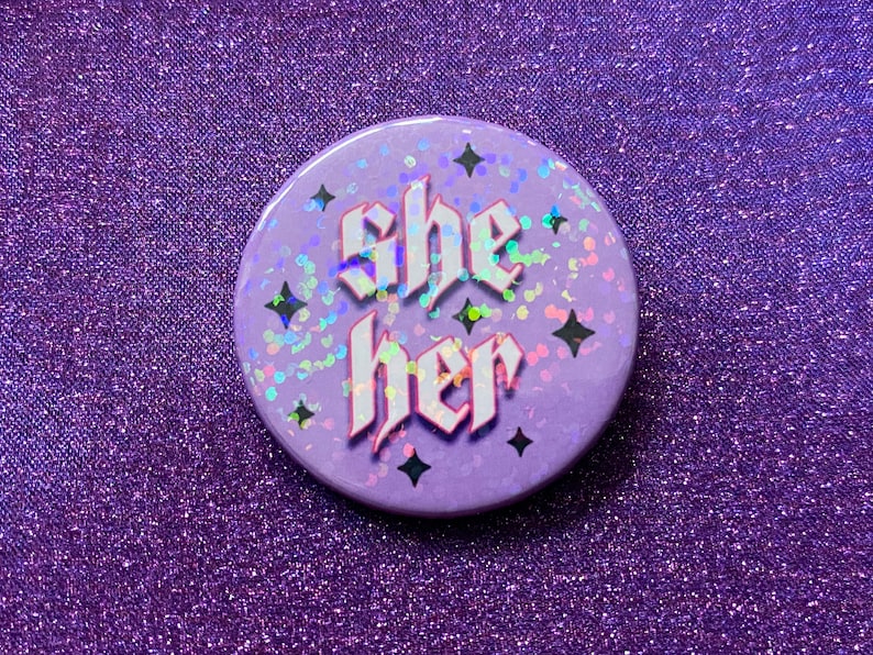 May include: A purple button with a glittery surface and black stars. The button says "she her" in white letters with a gothic font.