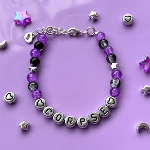 May include: A purple and black beaded bracelet with silver charm beads spelling out the word "CORPSE".