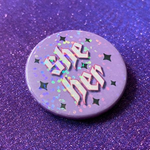 May include: A purple button with a holographic finish and black stars. The button says "she her" in white gothic lettering.