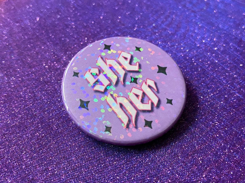 May include: A purple button with a holographic finish and black stars. The button says "she her" in white gothic lettering.