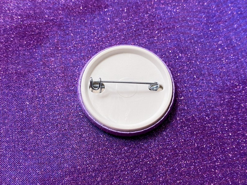 May include: The back of a purple button with a metal pin and clasp.