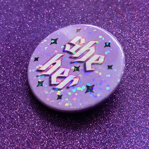 May include: A purple button with iridescent glitter and the words "She Her" in white with a pink outline.