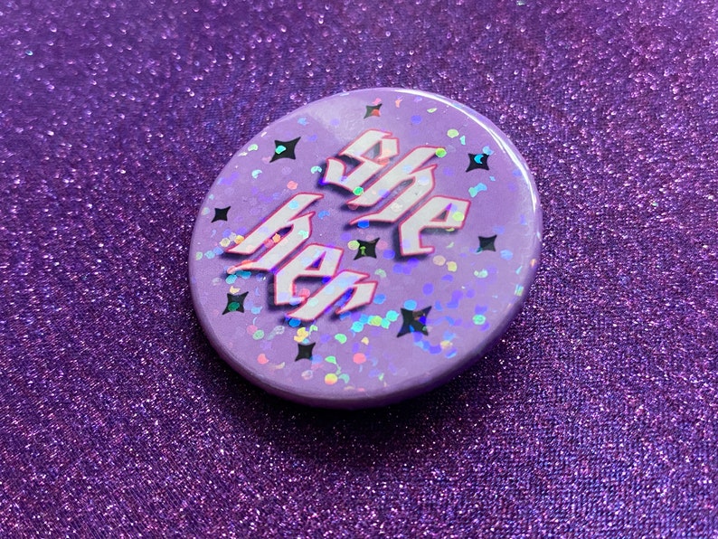 May include: A purple button with iridescent glitter and the words "She Her" in white with a pink outline.