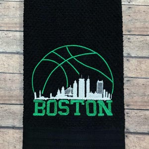 Puede incluir: Toalla de mano negra con un diseño de baloncesto verde y la palabra "BOSTON" bordada en verde. El diseño incluye una pelota de baloncesto y una silueta del horizonte.