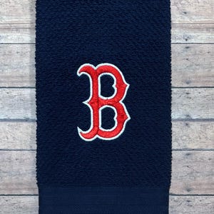 May include: A navy blue hand towel with a textured surface. It features a large, embroidered red and white "B" logo in the centre. The bottom of the towel has a solid navy blue band.