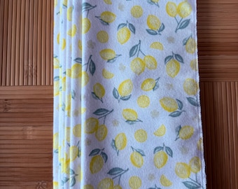 May include: A stack of six white dishcloths with a lemon print. The dishcloths are folded in half and have a yellow stitched edge.