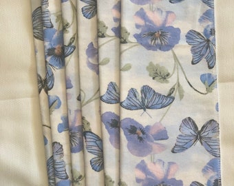 May include: A stack of folded white cloth napkins with a repeating pattern of blue butterflies and purple flowers. The napkins are arranged on a white surface, showcasing the floral and insect design.
