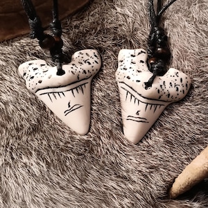 May include: Two off-white shark tooth pendants with black detailing hang from black cords. The teeth have a textured top and a detailed bottom edge. The pendants rest on a textured, gray and brown surface, with a portion of a light-colored antler in the background.