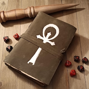 May include: A leather-bound journal with a white ankh symbol and a leather strap closure. A wooden wand and several dice are arranged around the journal on a wooden surface. The journal is olive green.
