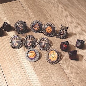 May include: A collection of nine circular and oval metal brooches featuring dark purple and orange graphic symbols under glass cabochons, accompanied by four dark purple multi-sided dice with white numbers scattered among the jewellery pieces.