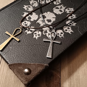 May include: Two ankh necklaces rest on a dark, textured surface decorated with skull motifs. One ankh is gold-toned, the other silver. The necklaces are displayed on a book with leather-like accents and metal studs, set against a wooden background.