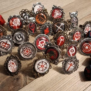 Könnte beinhalten: Eine Sammlung verzierter Ringe mit verschiedenen roten, weißen und schwarzen Designs, zusammen mit roten und schwarzen Würfeln. Die Ringe haben filigrane Fassungen und zeigen Symbole, Kronen und geometrische Muster.
