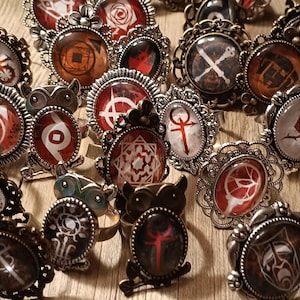 May include: A collection of assorted rings featuring ornate metal settings and glass cabochons with various graphic designs, including symbols, roses, and owl motifs in shades of red, black, white, and bronze, arranged in a group on a flat surface.