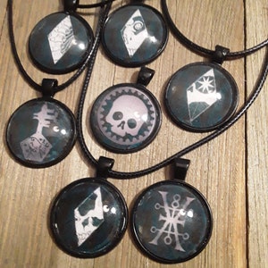 Könnte beinhalten: Sieben schwarze Halsketten mit runden Anhängern, die mit weißen grafischen Designs verziert sind. Die Anhänger haben einen dunkelblauen Hintergrund mit einem weißen Rand. Die Designs beinhalten einen Schädel, einen Stern, einen Diamanten und ein geometrisches Muster.