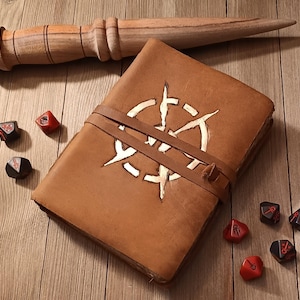 May include: A brown leather journal with a white compass design and a leather strap closure. A wooden wand and several red and black dice are arranged around the journal on a wooden surface. The journal measures approximately 8 inches by 6 inches.