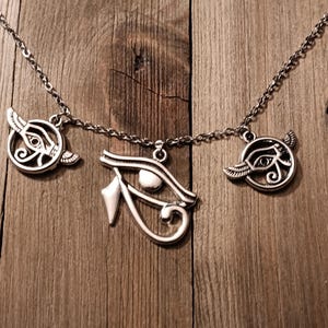 May include: Silver-toned necklace featuring three pendants with Egyptian Eye of Horus designs. The pendants are suspended from a delicate chain, set against a wooden background. The necklace is a piece of jewelry.
