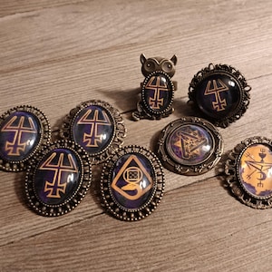 May include: An assortment of antique-style rings and brooches with mystical symbols. The jewelry features dark blue and orange designs set in ornate, bronze-colored metal frames. The collection includes oval and round shapes, with one ring shaped like an owl.