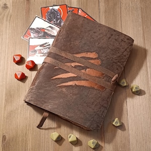 May include: A dark brown leather journal with a textured surface and a prominent claw mark design on the cover, secured by a leather strap. The journal is surrounded by several small, multi-sided gaming dice in red and light green, along with a fan of illustrated cards featuring dark, creature-themed artwork.
