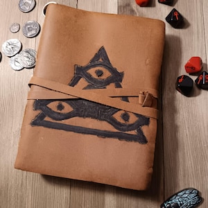 May include: A brown leather journal with a black eye symbol on the front. The journal has a strap closure and is surrounded by coins and dice.