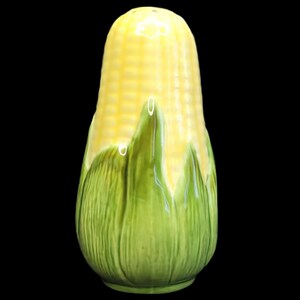Puede incluir: Un salero de cerámica con forma de mazorca de maíz con un acabado amarillo brillante en los granos y hojas verdes texturizadas en la base. El objeto presenta una parte superior redondeada y lisa y una forma cilíndrica con detalles.