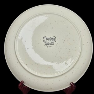 May include: White ceramic plate with a brown speckled glaze. The bottom of the plate has a black and white label that reads "Fantasy Santa Ana Ware Made in California Trademarked for Rose-ware Copyright".