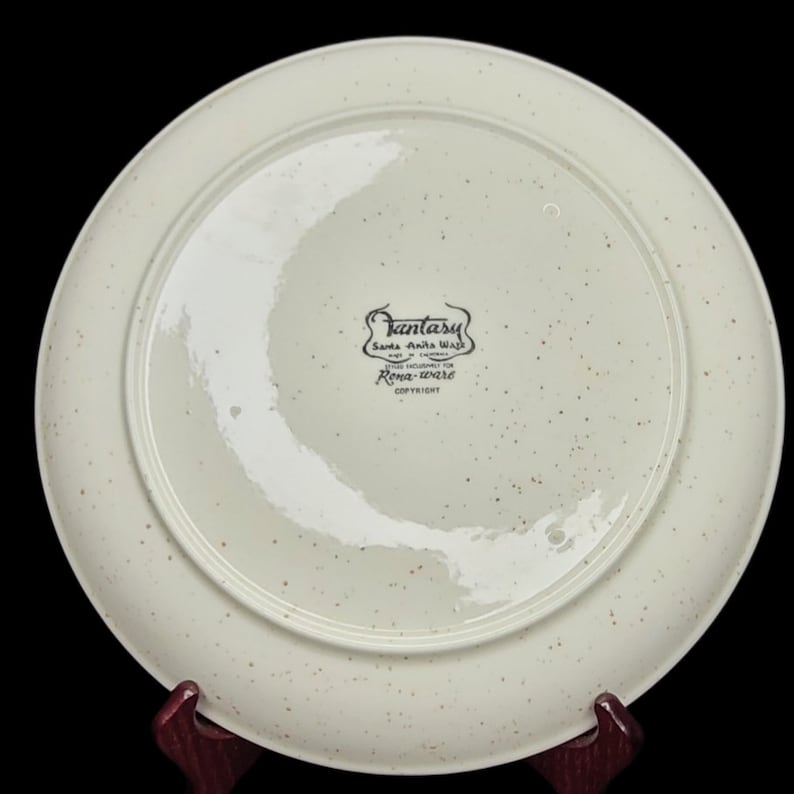 May include: White ceramic plate with a brown speckled glaze. The bottom of the plate has a black and white label that reads "Fantasy Santa Ana Ware Made in California Trademarked for Rose-ware Copyright".