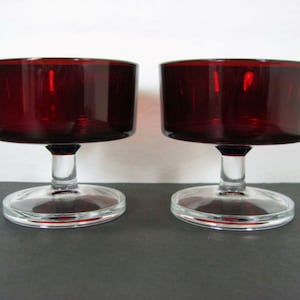 Può includere: Due coppe da dessert in vetro trasparente con gambo e una ciotola rosso scuro. Le coppe sono vuote e sono su una superficie bianca.