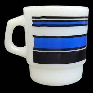 Puede incluir: Taza de cerámica blanca con un patrón geométrico azul y negro. El patrón presenta tres rayas horizontales, con las rayas superior e inferior negras y la raya del medio azul.