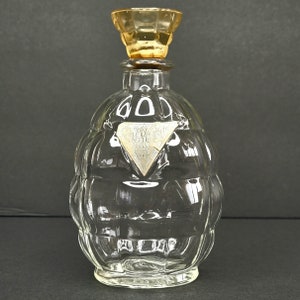 Puede incluir: Una botella de vidrio transparente con una superficie texturizada y una tapa de color dorado. La botella tiene una etiqueta en forma de diamante que dice "FLEUR D'INTERIEUR".