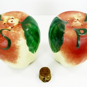 Op de afbeelding: Een set van twee keramische zout- en peperstrooiers in de vorm van rode appels met groene bladeren. De strooiers hebben gaten bovenop om zout en peper te strooien. Het woord "S" is op de ene strooier geschilderd en het woord "P" op de andere.