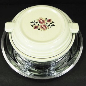 May include: A white ceramic serving dish with a floral design and a silver-plated base. The dish has a lid with a handle on each side.