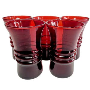 May include: A set of five deep red glass vases, each featuring a wide, flared rim and a series of three horizontal raised rings encircling the lower body. The glass is translucent, allowing light to pass through the vibrant crimson colour.